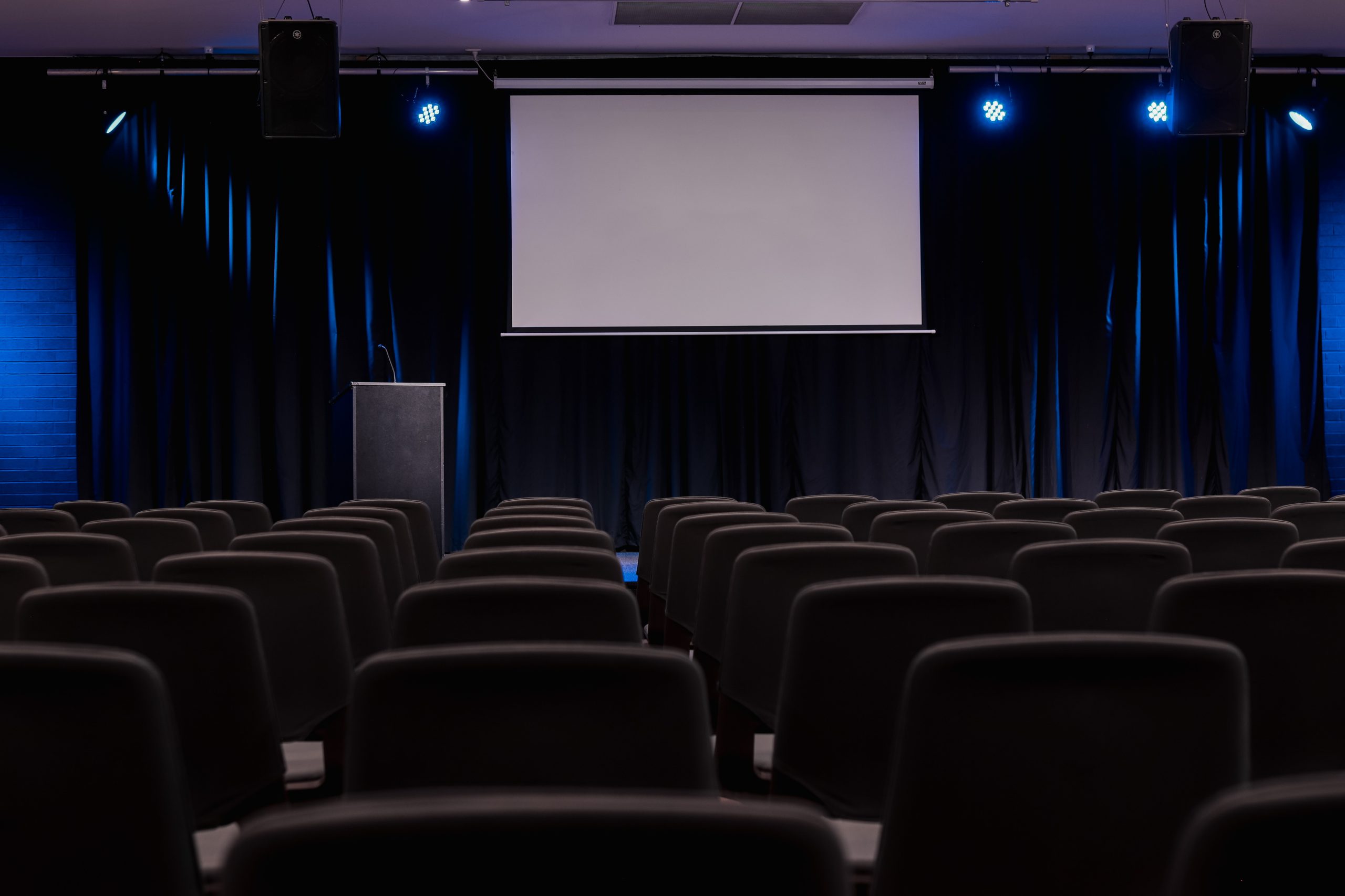 Group accommodation venue with auditorium. Blue stage lights shine, a projector on the wall and a podium on the stage.