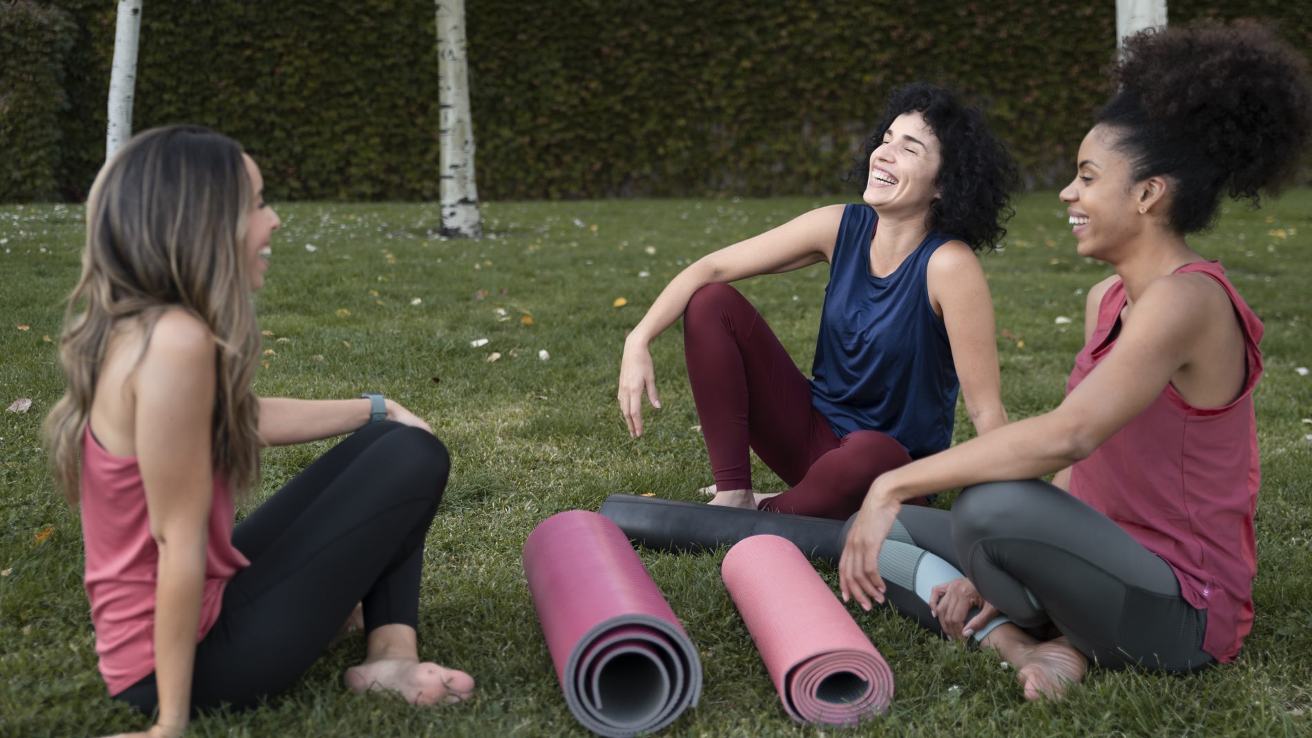Three women sit on the grass and laugh together at a fitness retreat.